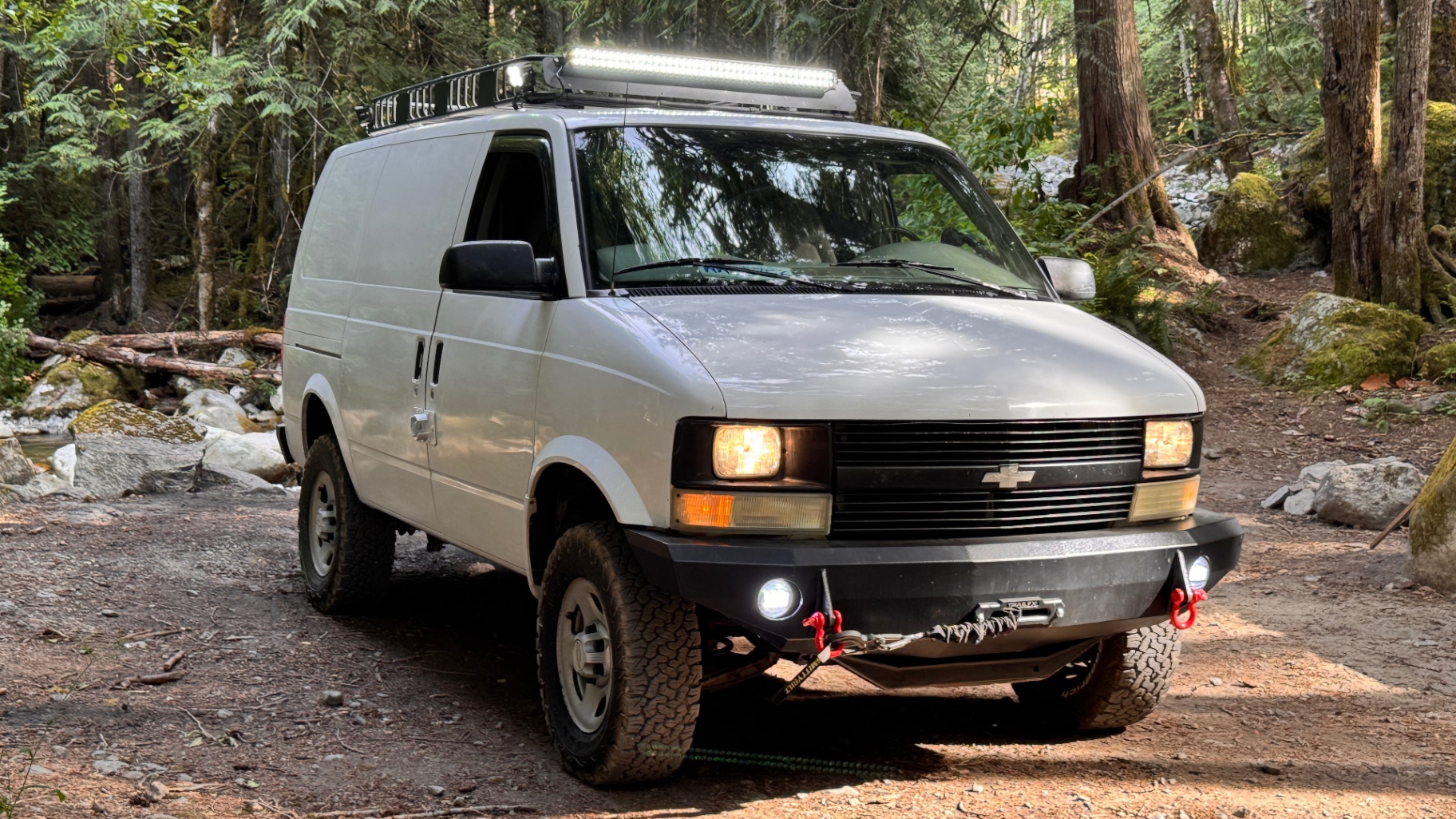 Three-quarter view of the Astro on a creekside FSR pullout, light bar mounted on the rack, JOR bumper and fog lights visible