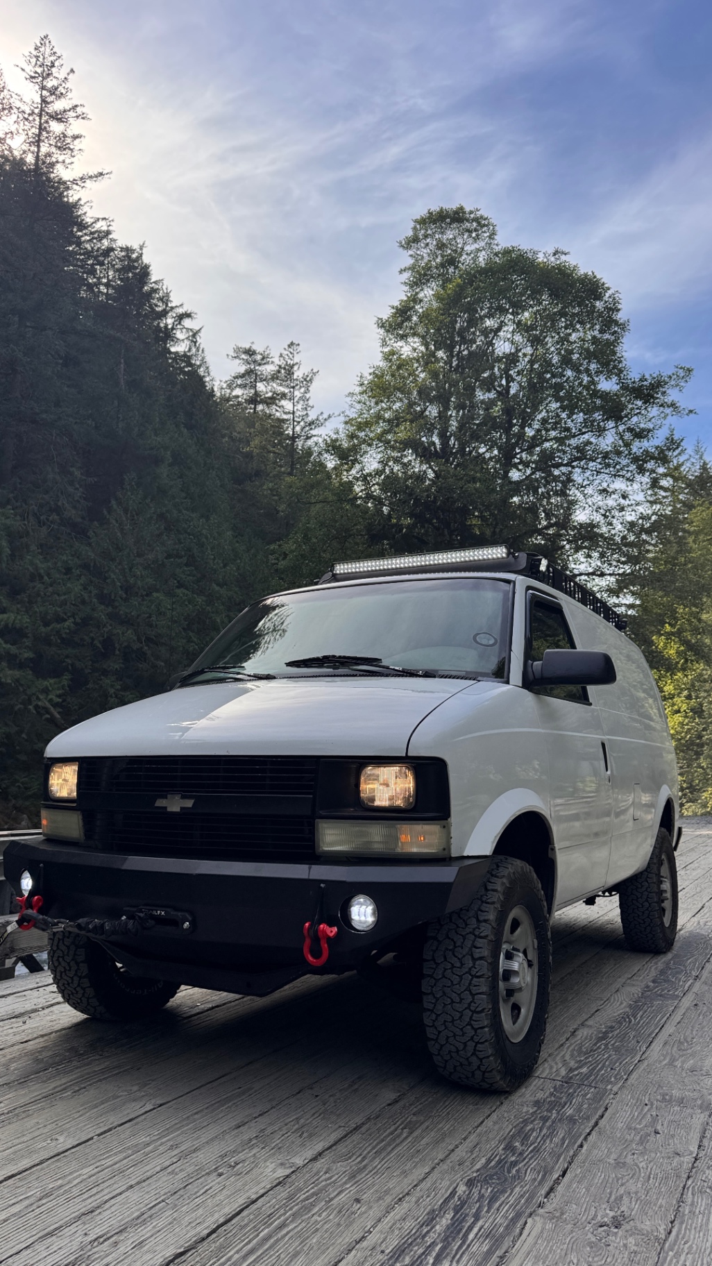 Astro parked on a wooden FSR bridge, low afternoon sun behind the cedars, JOR bumper with red recovery hooks and fog lights on